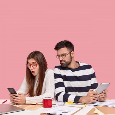 Team of young people brainstrom together, sit back to each other, addicted to modern technologies, use mobile phone and touchpad, pose at desktop with documentation, have briefing meeting, isolated