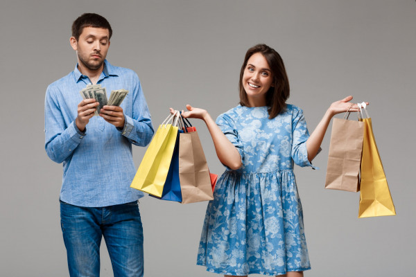 Young beautiful woman holding purchases, man holding money over grey background. Copy space.